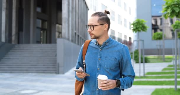 Kaukázusi fiatalember szemüvegben, aki mobilon tapogatózik, és kávét tart elvitelre a City Streeten. Jóképű férfi szemüvegben sms-t ír az okostelefonon a szabadban. Szerkentyű használata koncepció. — Stock videók