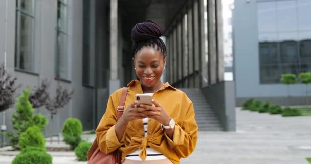 Afroamericana alegre joven elegante mujer tocando o desplazándose en el teléfono inteligente y de pie en la calle de la ciudad. Hermoso mensaje de texto femenino feliz en el teléfono móvil y sonriendo. Afuera.. — Vídeos de Stock
