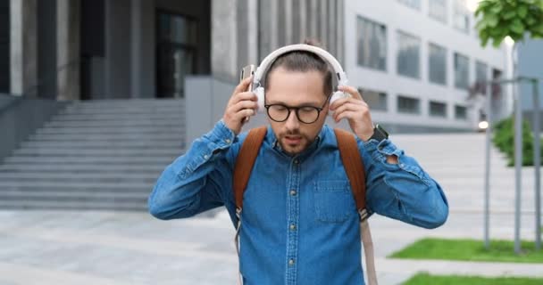 ヘッドフォンで撮影し、音楽を屋外で聞き、街の通りを歩くメガネの白人ハンサムな若い男。スマートフォンをタップして使用しています。男性学生バックパック付き. — ストック動画