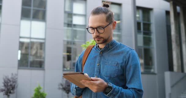 Retrato de un joven caucásico con gafas golpeando en un dispositivo de tableta y sonriendo a la cámara en la calle de la ciudad. Guapo mensaje de texto masculino y la navegación en la computadora gadget al aire libre. Gadget concepto de usuario. — Vídeos de Stock