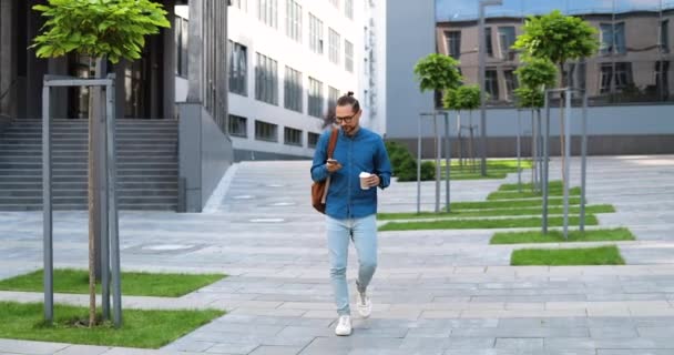 Un jeune homme caucasien descend la rue en tapant sur son smartphone et en sirotant un verre. Homme gooing bas de l'échelle en plein air, textos mesage sur le téléphone et boire du café à emporter. Se précipiter le matin. — Video