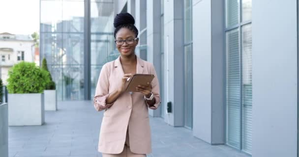 Afro-Amerikaanse vrolijke jonge stijlvolle zakenvrouw in glazen lopen buiten en met behulp van tablet-apparaat. Mooie glimlachte vrouw tikken en scrollen op gadget computer tijdens een wandeling op straat. — Stockvideo