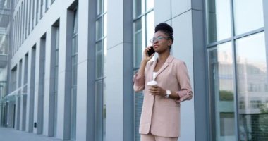 Gözlüklü Afro-Amerikalı genç bir kadın iş merkezinde cep telefonuyla konuşuyor. Gözlüklü kendine güvenen bir iş kadını cep telefonuyla konuşuyor ve elinde bir fincan kahve tutuyor..