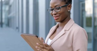 Afro-Amerikalı genç ve gülümseyen gözlüklü iş kadınını yakından takip edin. Tablet cihazına tıklayıp kaydırın. Dışarıdaki bilgisayardan mesaj yollayan bir kadın. Kendine güveni tam bir kadın, alet kullanıyor ve konuşuyor..