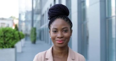 Pembe ceketli güzel Afro-Amerikan bir kadının portresi. Kameraya bakıyor ve dışarıda neşeyle gülümsüyor. Çekici, mutlu, gülümseyen iş kadını iş merkezinin yanındaki sokakta..