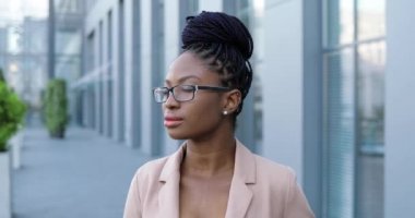 Pembe ceketli, gözlüklü, yüzü kameraya dönük ve neşeli bir şekilde gülümseyen genç Afro-Amerikan kadın portresi. İş merkezinin yakınındaki caddede çekici, neşeli bir iş kadını..