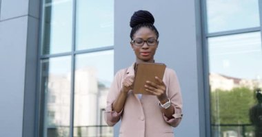 Gözlüklü Afro-Amerikalı genç iş kadını tablet cihazını tıklatıyor ve kaydırıyor. Dışarıdaki bilgisayardan mesaj yollayan bir kadın. Kendine güveni tam bir kadın, alet kullanıyor ve konuşuyor..