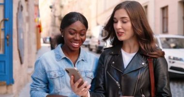 Beyaz ve Afrikalı Amerikalı bayan arkadaşlar caddede dikilip konuşuyorlar ve cep telefonundan bir şeyler izliyorlar. Çok ırklı güzel kadınlar sohbet ediyor ve akıllı telefon kullanıyor. Dedikoducu kızlar. Dedikodular