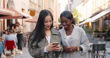 Caddede yürüyen, konuşan ve telefonda video izleyen melez kadınlar. Çok ırklı genç kadınlar sohbet ediyor ve akıllı telefon kullanıyor. Neşeli Asyalı ve Afrikalı Amerikalı kızlar dedikodu yapıyor. Dedikodular