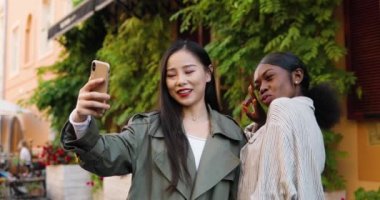 Neşeli Asyalı ve Afro-Amerikalı güzel kadınlar akıllı telefon kamerasına poz veriyor ve gülümsüyor. Karışık ırktan genç kadınlar telefonla selfie çekiyorlar. Çok ırklı kızlar dışarıda fotoğraf çekiyor..