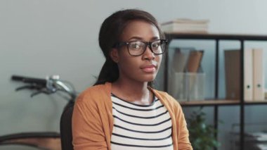 Modern ofiste oturmuş gülümseyen kameraya bakarken gözlüklü ve sıradan giyinmiş Afro-Amerikan iş kadınının portresini yakından çek..