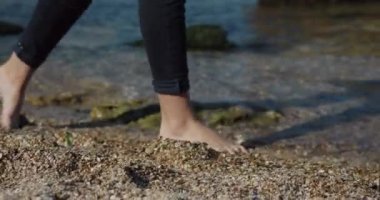 Kamerayı kapat ve kot pantolonlu bacakları takip et. Dişi bacaklar deniz sahilinde yalınayak yürür. Kıyı şeridi. Turistik yer.