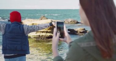 Havalı kıyafetler içindeki güzel modern kız, arkadaşı akıllı telefonuyla fotoğraf çekerken deniz kıyısında dikilirken kollarını açıyor..