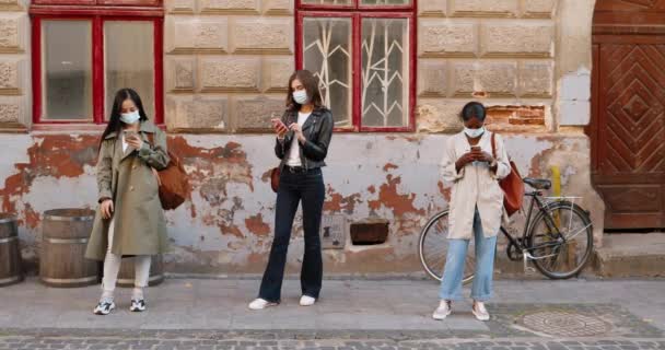 Bastante mestizas hembras en máscaras médicas de pie al aire libre y mensajes de texto en los teléfonos inteligentes. Distancia social. Mujeres jóvenes multiétnicas tocando y desplazándose en teléfonos móviles. Línea de espera. — Vídeos de Stock