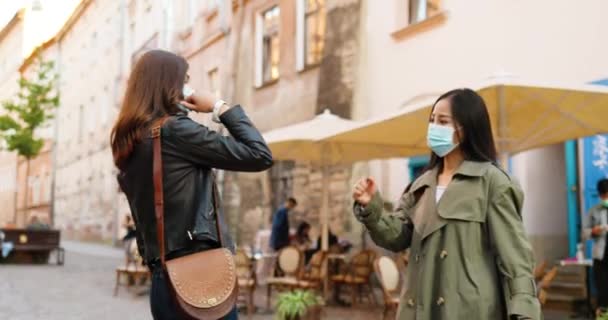 Blandade raser unga kvinnor, bästa vänner möte på stadens gata och hälsning med armbågar under coronavirus pandemi. Multietniska kvinnor i medicinska masker pratar utomhus. Kaukasiska och asiatiska flickor. — Stockvideo