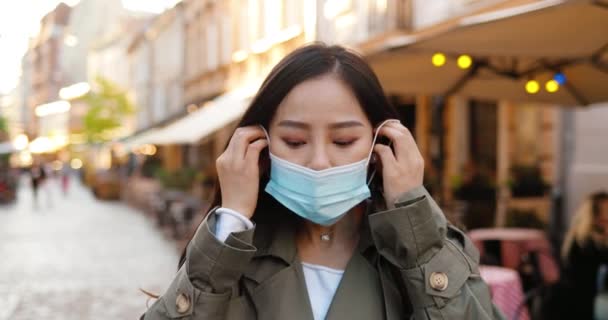 Portrait of beautiful young stylish Asian woman looking at camera and wearing medical mask at street. Pretty female taking on respiratory protection outdoor. Pandemic concept. Coronavirus. Close up. — Stock Video