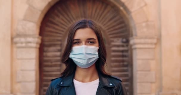 Portrait of beautiful young Caucasian woman in black leather jacket and medical mask looking at camera cheerfully at street. Outdoor. Close up of female face in respiratory protection Pandemic concept — Stock Video