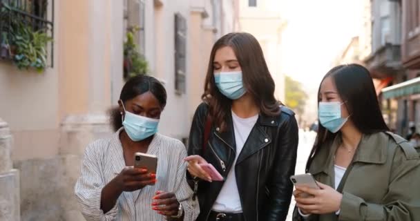 Drie gemengde rassen jonge stijlvolle vrouwen in medische maskers praten en lachen tijdens het lopen op straat en met behulp van smartphones. vrij multi etnische vrouwen vrienden wandelen buiten en afluisteren op telefoons. — Stockvideo