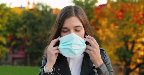 Retrato de una hermosa joven caucásica mirando a la cámara y usando máscara médica en el parque. Bonita hembra tomando protección respiratoria afuera. Concepto de medidas seguras pandémicas. Coronavirus — Vídeos de Stock