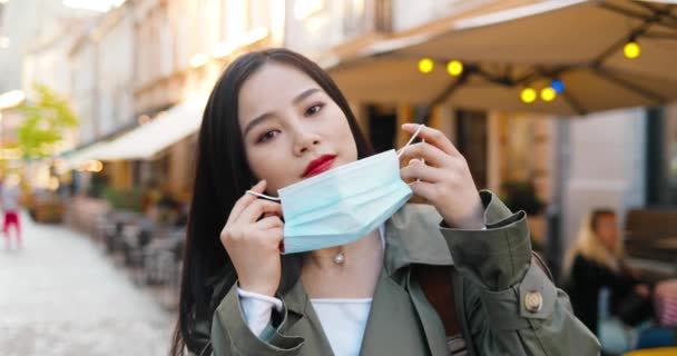 Portré gyönyörű fiatal, stílusos ázsiai nő néz kamera és visel orvosi maszkot az utcán. Közel egy csinos nőhöz, aki lélegeztetőgéppel védekezik a szabadban. Pandémiás fogalom. Koronavírus — Stock videók