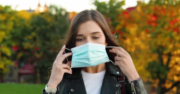 Retrato de bela jovem mulher branca elegante olhando para a câmera e vestindo máscara médica no parque. Uma mulher bonita a receber protecção respiratória ao ar livre. Conceito de medidas pandémicas. Coronavírus. — Vídeo de Stock