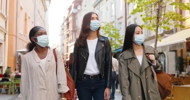 Trzy mieszane rasy młodych stylowych kobiet w maskach medycznych mówiących i spacerujących ulicą w mieście. Całkiem wesołe wielonarodowe przyjaciółki spacerujące na świeżym powietrzu i oglądające miasto. Turyści w czasie pandemii. — Wideo stockowe