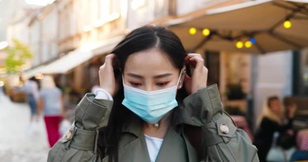 젊고 아름다운 아시아 여성이 카메라를 보고 있고 길거리에서 의료용 마스크를 쓰고 있는 모습. 아름다운 암컷이 바깥 도시에서 호흡기 보호를 받고 있습니다. 세계적 인 컨셉이야. Covid-19. — 비디오