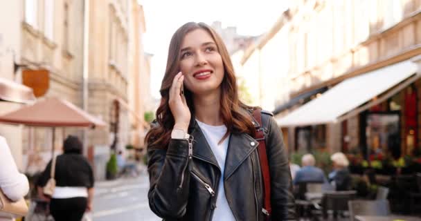 Красивая кавказская молодая стильная женщина в черной кожаной куртке, улыбающаяся и разговаривающая по мобильному телефону во время прогулки по улице. Красивая счастливая веселая женщина говорит по телефону и гуляет по городу. — стоковое видео