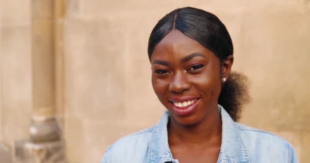 Портрет гарної афроамериканської щасливої молодої жінки, яка дивиться і посміхається фотоапарату на стіні. На вулиці. Зображення прекрасної веселої жіночої посмішки. — стокове відео