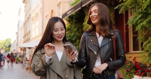 Mezclas de razas femeninas amigas caminando por la calle, hablando y viendo vídeo en el teléfono móvil. Multi étnicas mujeres jóvenes charlando y utilizando el teléfono inteligente. Alegre Asiático y Caucásico niñas chismorreando. Chismes — Vídeos de Stock