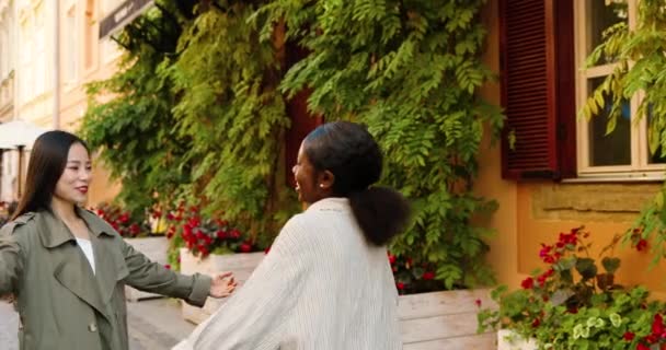 Jóvenes mestizos hermosas mujeres alegres que se reúnen en la calle y se abrazan felizmente. Mujeres bonitas multiétnicas en abrazos riendo y hablando. Al aire libre. Afro Americano y Asiático niñas mejores amigos abrazar — Vídeo de stock