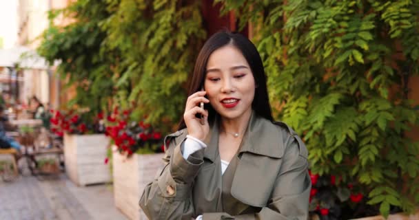 Jolie asiatique jeune femme élégante souriant et parlant sur téléphone mobile sur fond de mur de plantes vertes. En plein air. Belle femme heureuse et joyeuse parlant sur son téléphone portable. Jolie conversation. Appel téléphonique. — Video