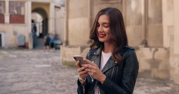 Bella giovane donna caucasica elegante in giacca di pelle nera sorridente e messaggio di testo sul telefono cellulare in strada. Bella felice allegra femmina toccando e scorrendo su smartphone in città. — Video Stock