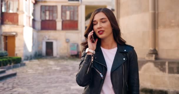 Wanita muda bergaya Kaukasia cantik dengan jaket kulit hitam tersenyum dan berbicara di telepon genggam saat berjalan di jalan. Indah bahagia perempuan ceria berbicara di ponsel dan berjalan-jalan di luar ruangan. — Stok Video
