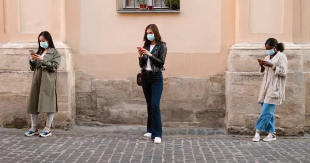 Dość mieszane rasy kobiet w maskach medycznych stojących na zewnątrz i wiadomości tekstowych na smartfonach. Odległość społeczna. Młode, wieloetniczne kobiety podsłuchujące i przewijające telefony komórkowe w kolejce. — Wideo stockowe