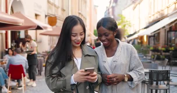 Mixed-races females friends walking the street, talking and watching video on phone. Multi ethnic young women chatting and using smartphone. Joyful Asian and African American girls gossiping. Gossips — Stock Video