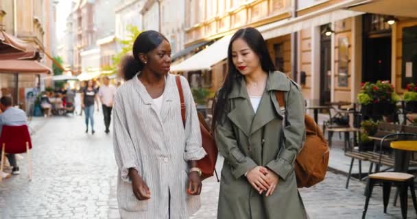 Mixed-races females best friends walking the street, talking and sharing secrets. Multi ethnic young women chatting and strolling outside. Cheerful Asian and African American girls gossiping. Gossips. — Stock Video