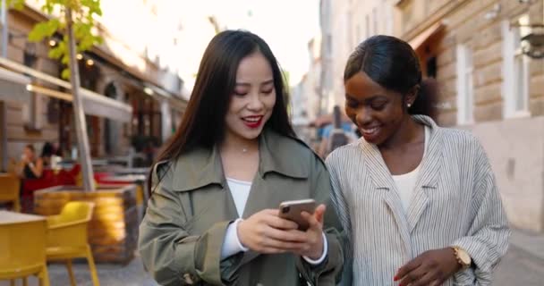 Смешанные гонки женщины ходят по улице, разговаривают и смотрят видео по телефону. Молодые женщины разных национальностей общаются и пользуются смартфонами. Веселая азиатка и афроамериканка сплетничают. Гемпс. — стоковое видео