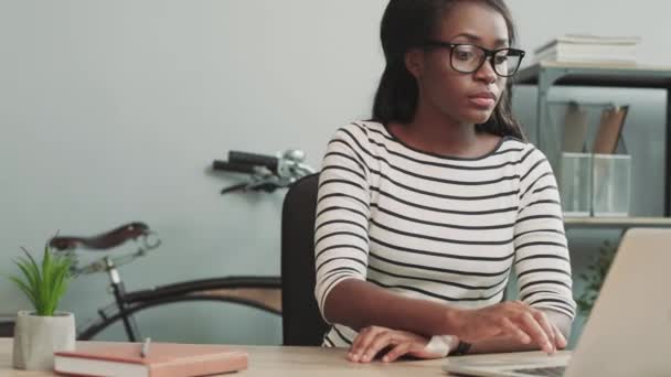 Młoda piękna dziewczyna siedzi w biurze przy biurku przegląda dane na swoim laptopie i wprowadza je do inteligentnego zegarka. — Wideo stockowe