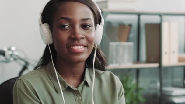 若いです美しいアフリカ系アメリカ人の女の子は白いヘッドフォンで音楽を聞き、オフィスに座っている間に良い音を楽しみます. — ストック動画