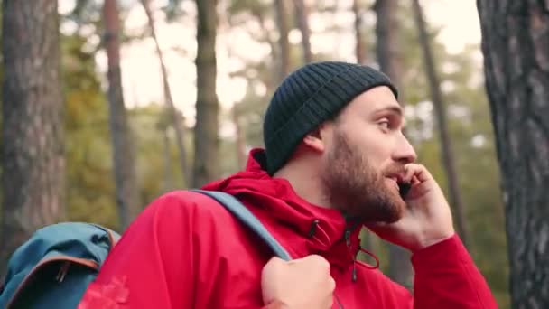 Fiatal, elegáns fehér férfi szakállal áll az erdő közepén hátizsákkal, és beszél a mobiltelefon barátaival mosolyog. — Stock videók