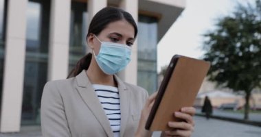 Tıp maskeli güzel kadın işçinin portresi şehrin dışında duruyor ve tablet üzerinde daktilo yazıyor. Kafkasyalı genç bir kadın cihaza dokunuyor ve araştırıyor. Ağır çekim. İş konsepti