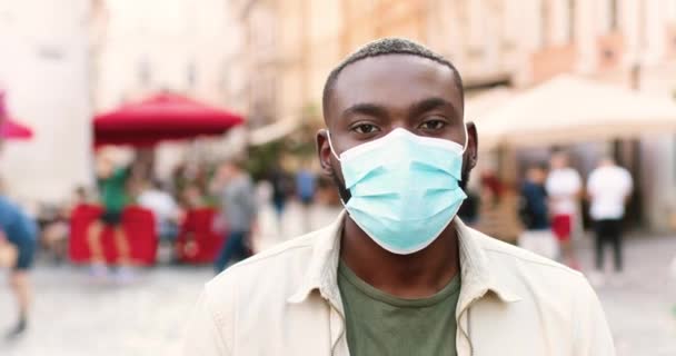 Retrato de un joven afroamericano guapo con máscara médica parado en la calle y mirando a la cámara. Concepto pandémico. Primer plano de la cara masculina en protección respiratoria. Medidas de cuarentena. — Vídeos de Stock