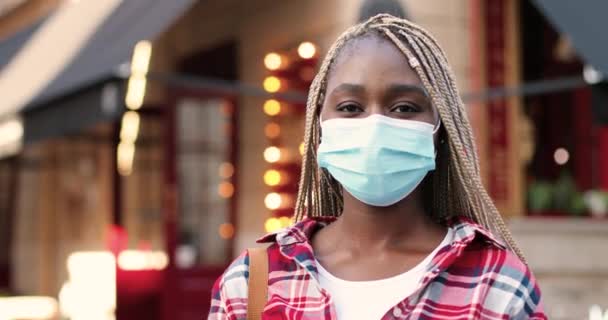 Retrato filmado de afro-americano jovem mulher elegante em máscara médica olhando para a câmera e de pé na rua perto do café. Close up de bela fêmea com tranças ao ar livre. Conceito de corona pandêmica. — Vídeo de Stock