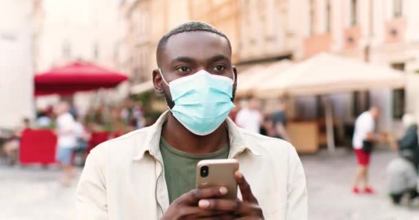 Закрийте молодого афроамериканського стиліста в медичній масці, який стоїть на вулиці і надсилає смартфон. Поручні самець стукає і прокручує по мобільному телефону.. — стокове відео