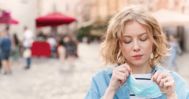 Close up of Caucasian young beautiful woman taking on medical mask and looking at camera at street in city. Portrait of pretty female outdoor wearing respiratory protection. Pandemic concept. — Stock Video