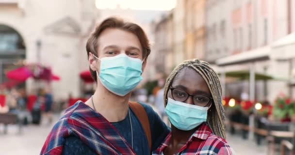 Portret młodej szczęśliwej mieszanej pary w maskach medycznych stojącej na zewnątrz ulicy i patrzącej w kamerę. Wielu etnicznych mężczyzn i kobiet w ochronie dróg oddechowych pozowanie do kamery w mieście. — Wideo stockowe