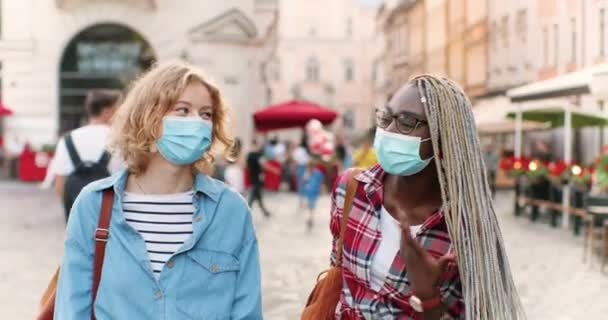 Two mixed-races cheerful females best friends in medical masks walking in city street and gossiping while having fun together. Young multi ethnic women smiling and talking when meet outdoor. — Stock Video