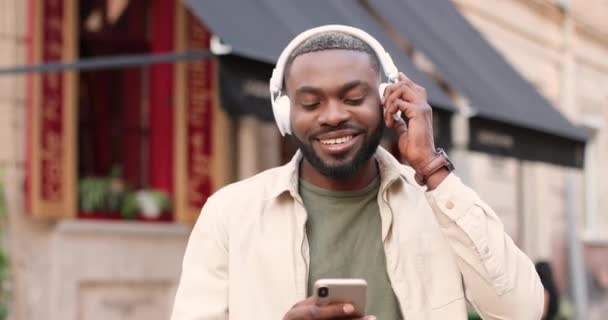 Joven afroamericano elegante hombre en grandes auriculares blancos caminando por la calle de la ciudad y escuchando música en el teléfono inteligente. Guapo feliz macho golpeando y desplazándose en el teléfono móvil y paseando. — Vídeos de Stock