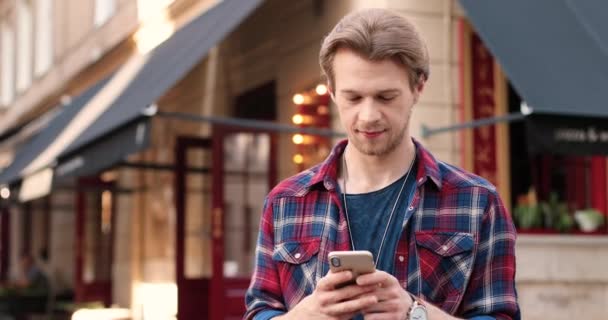 Молодий кавказький стиліст стоїть на вулиці біля кафе і пише смартфони. Поручні чоловічі телефони та прокрутки на мобільному телефоні. На вулиці. Використання мобільного телефону для обміну повідомленнями. — стокове відео